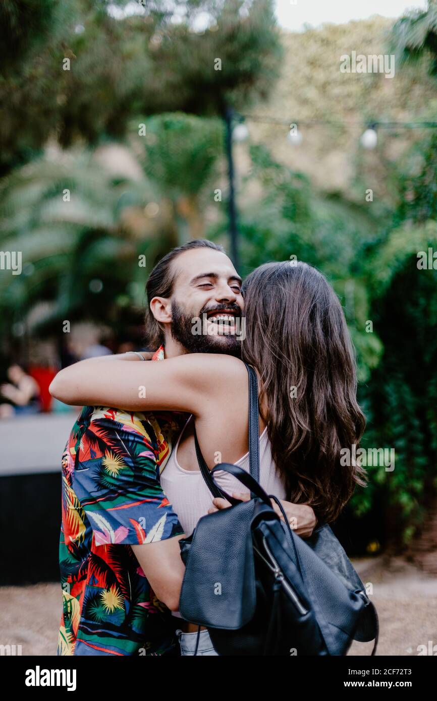 Vista lateral de un hombre con barba y una morena con mochila abrazar con los ojos cerrados sonriendo y de pie mientras abrazaba a los jóvenes morena contra el fondo de las plantas Foto de stock