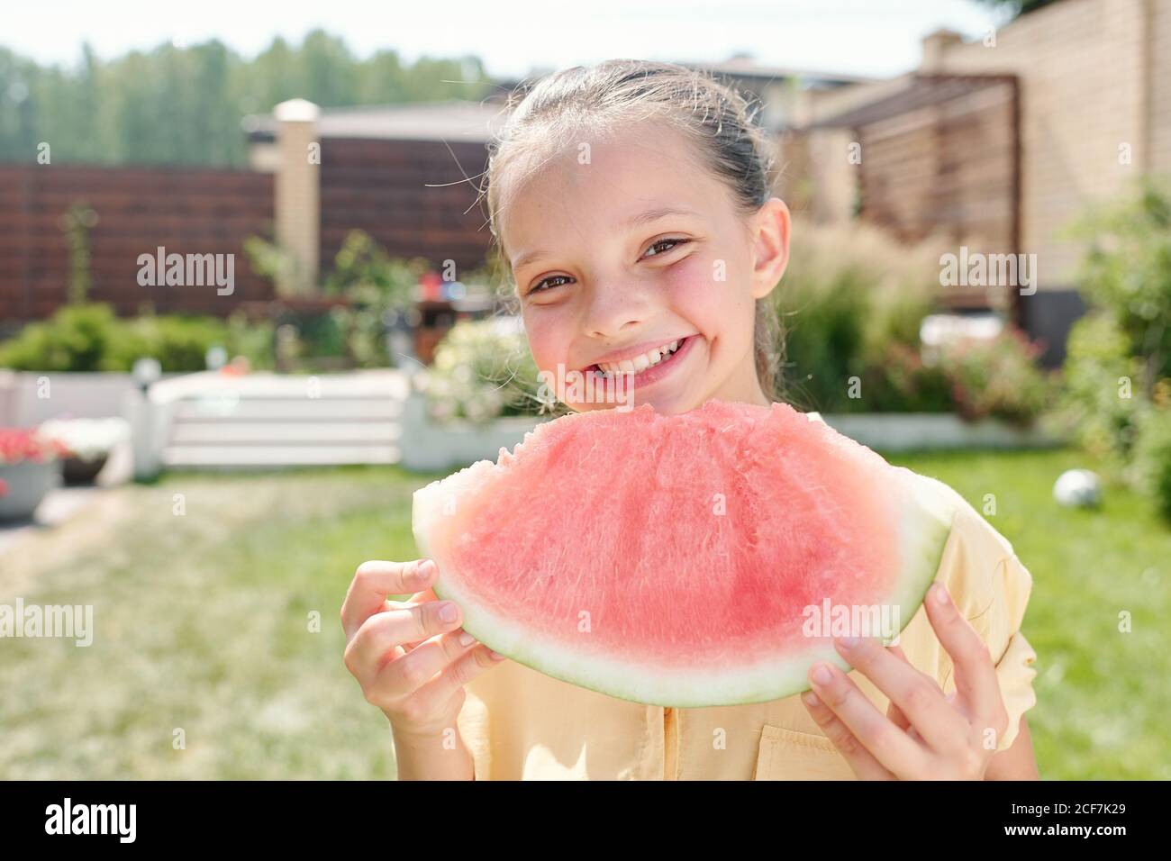 Retrato mediano de una niña feliz de pie en el patio trasero el día de verano sosteniendo una pieza de sandía sonriendo con la cámara Foto de stock