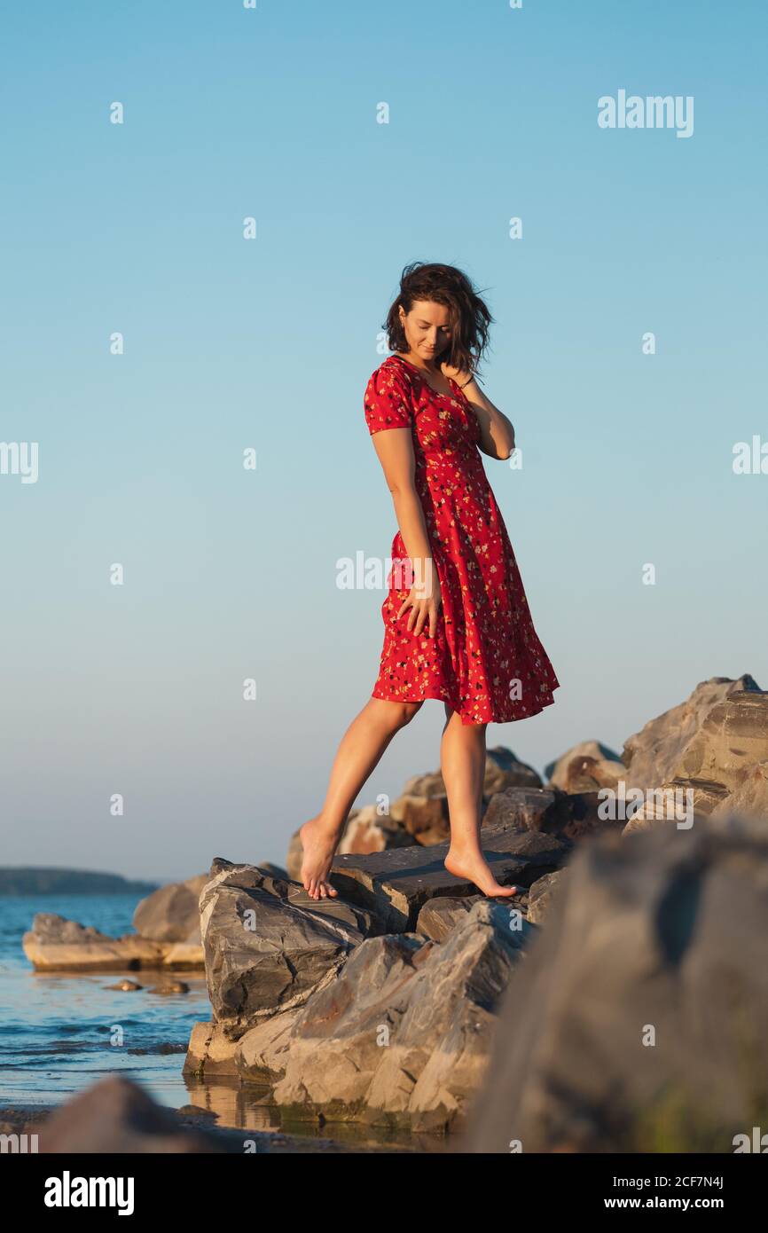 Joven hermosa mujer un vestido rojo informal ligero descansando y en las rocas en una playa de arena cerca de un cielo azul en un día verano, hermoso mar