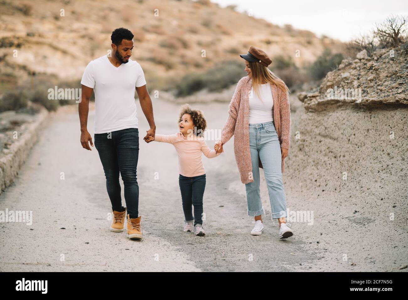 Feliz madre padre diverso y niño rizado con brillo casual ropa paseando por la naturaleza con las manos durante el día Foto de stock