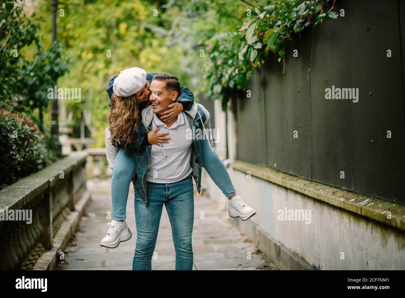 Mujer contenta con ropa informal sentada en la espalda y besándose feliz hombre caminando en un pequeño callejón con plantas verdes fondo borroso Foto de stock