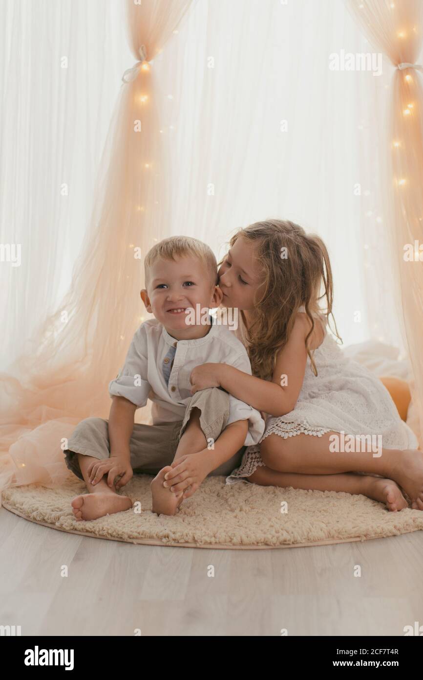 Niña con vestido blanco sentada en una alfombra abrace con un niño alegre y besando en la mejilla habitación elegante Foto de stock
