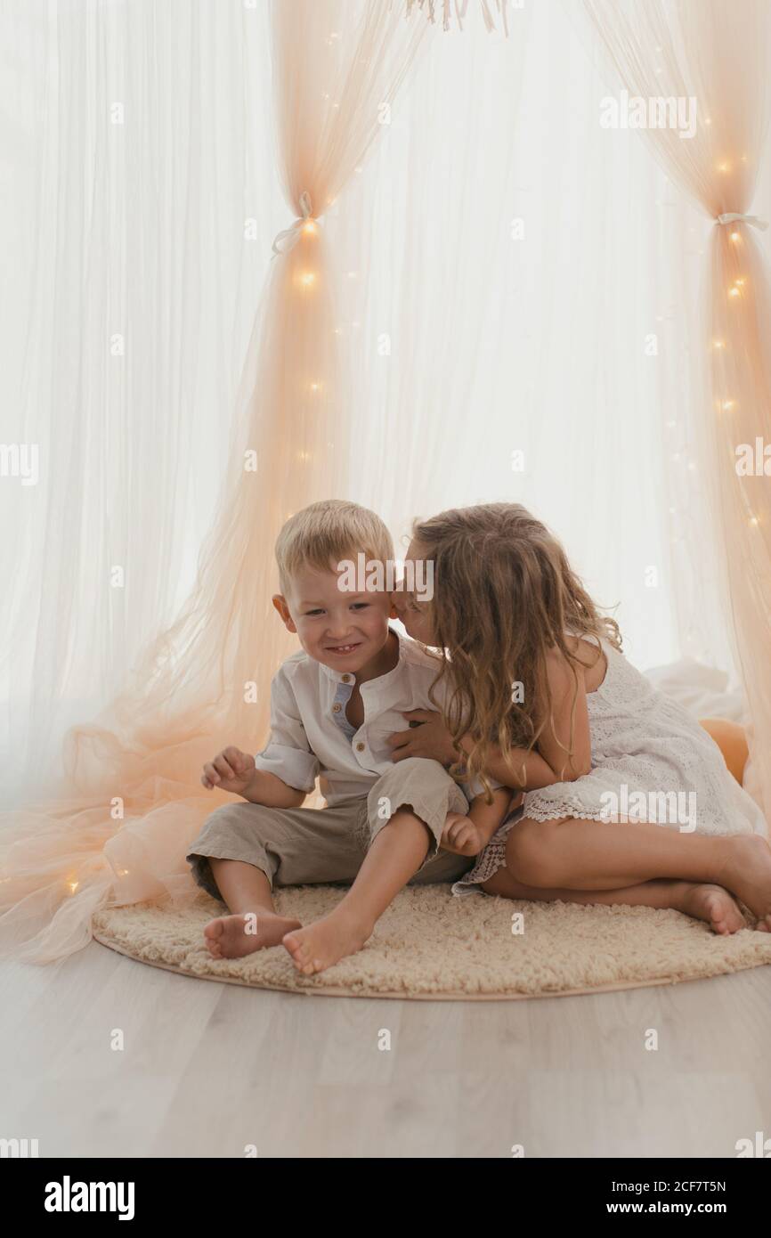 Niña con vestido blanco sentada en una alfombra abrace con un niño alegre y besando en la mejilla habitación elegante Foto de stock