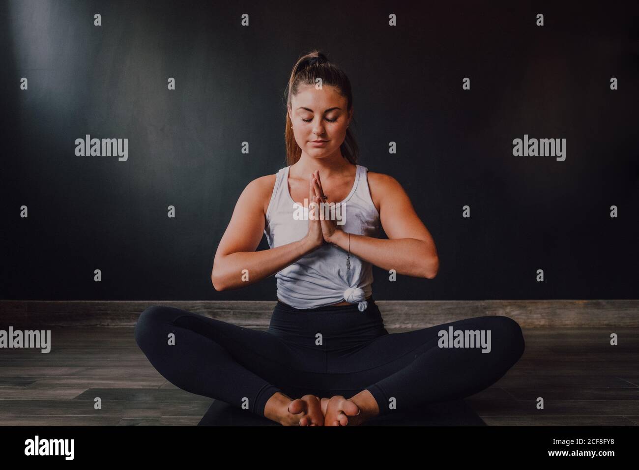 Mujer tranquila y relajada con los ojos cerrados y namaste sentado baddha konasana posición en la alfombra deportiva y concentrarse mientras practica yoga en estudio moderno oscuro Foto de stock