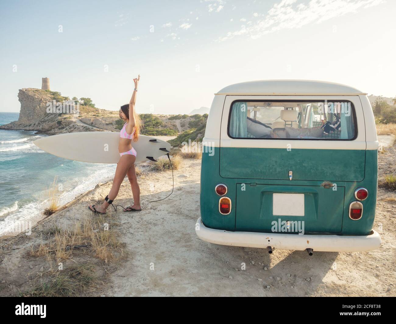Encantadora mujer de pelo largo que va a surfear sosteniendo una tabla de surf cerca de una furgoneta clásica Foto de stock