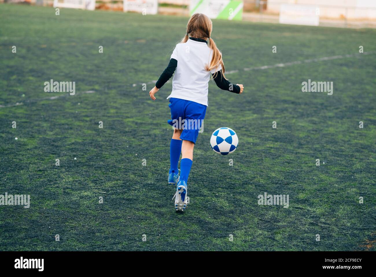 Vista posterior adolescente en blanco y azul uniforme y. tacos para correr  y prepararse para patear el balón mientras se juega al fútbol solo en el  campo verde en el club deportivo