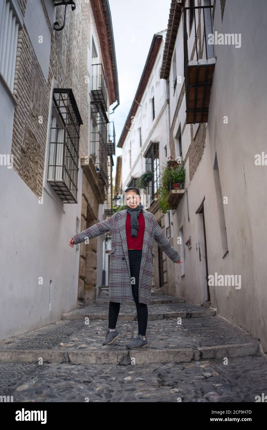 Desde abajo de la mujer viajera con alegría en ropa informal y.. elegante  abrigo sonriendo y de pie mientras levanta las manos y mira En la cámara  entre el carril en el