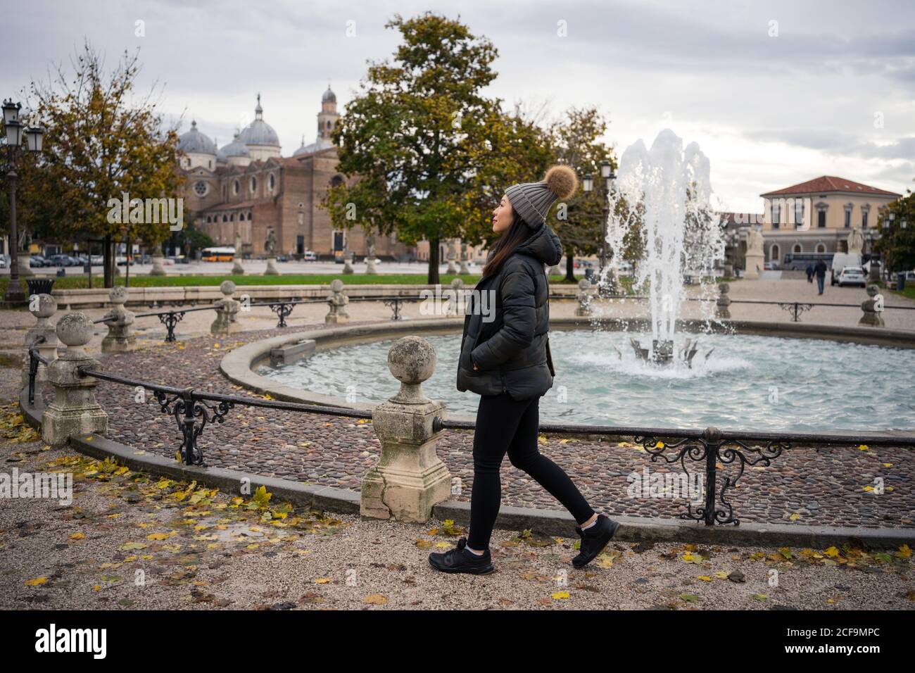 Vista lateral de la mujer viajera de pelo negro con ropa de abrigo Y  sombrero caminando en el hermoso parque antiguo Prato della Valle En Padova  en Italia Fotografía de stock -