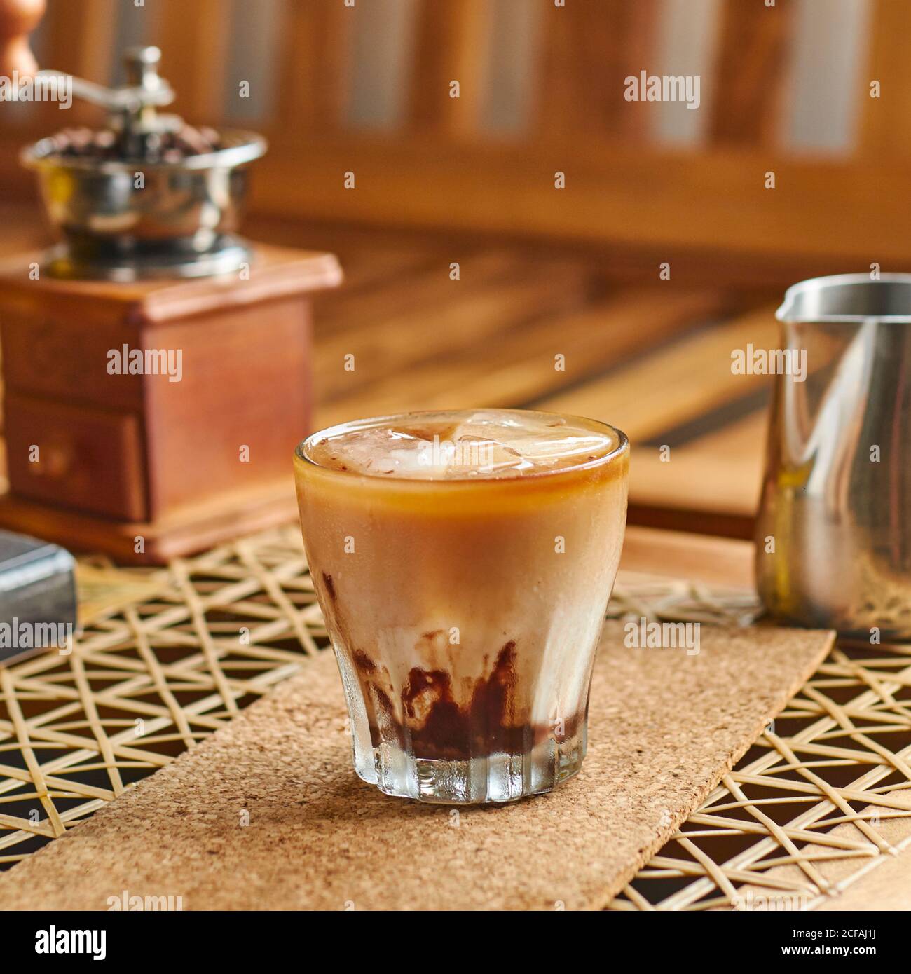 Gran ángulo de cristal transparente de delicioso café con fresco leche y  hielo en la montaña rusa de corcho junto al pequeño metal cafetera y  molinillo vintage sobre mesa de madera marrón