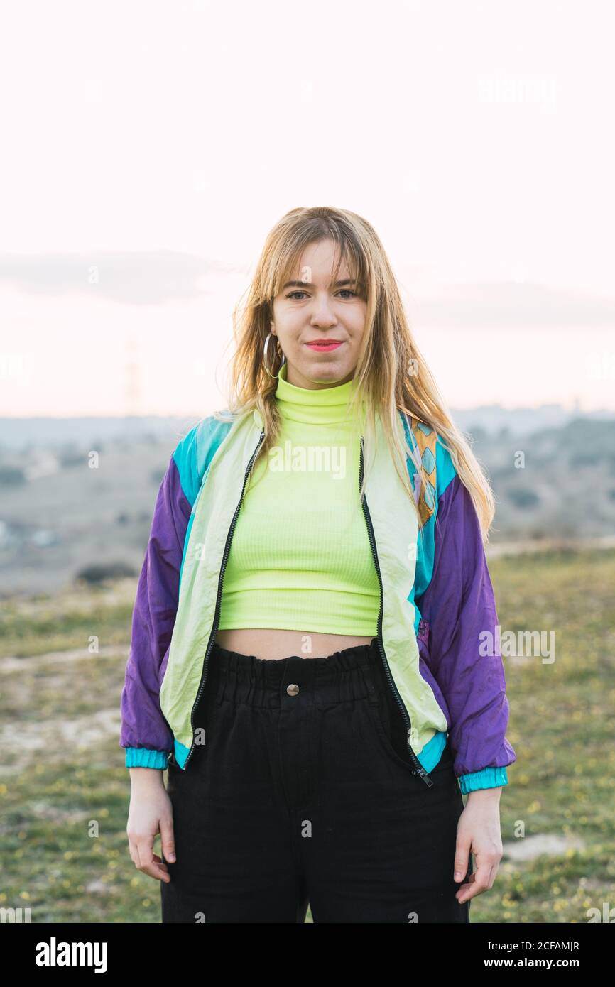 Mujer joven de pelo rubio alegre contemporánea en chaqueta de colores y..  parte superior de neón verde mirando a la cámara con la tierra remota  antecedentes Fotografía de stock - Alamy