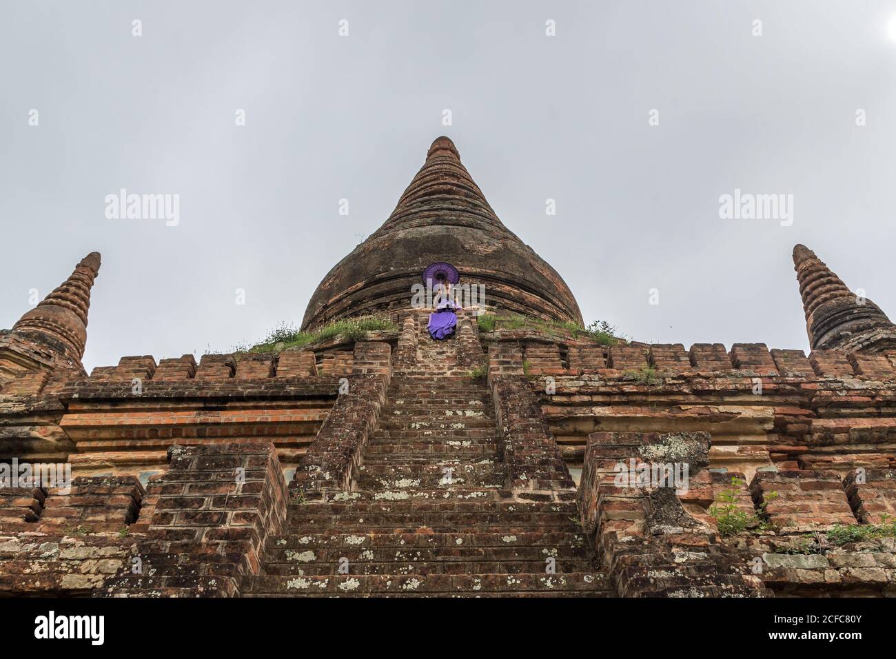 Desde abajo del antiguo templo de piedra birmano en Bagan contra gris cielo nublado con mujer en vestido púrpura sosteniendo púrpura paraguas en las escaleras Foto de stock