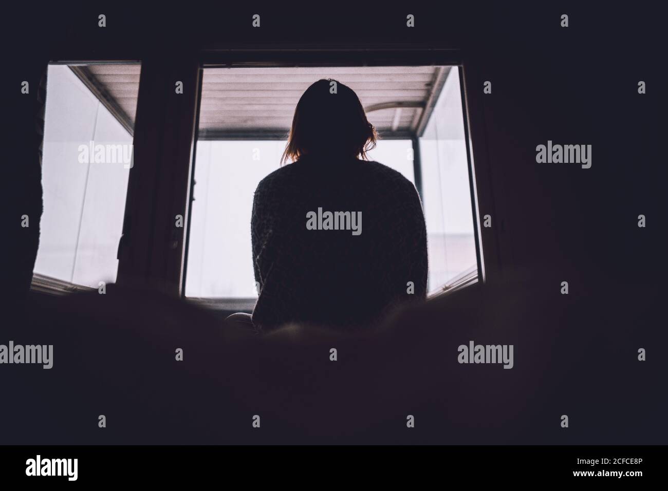 De la silueta del fuelle de la mujer solitaria anónima sentada en la cama contra la ventana en la habitación oscura en el hogar Foto de stock