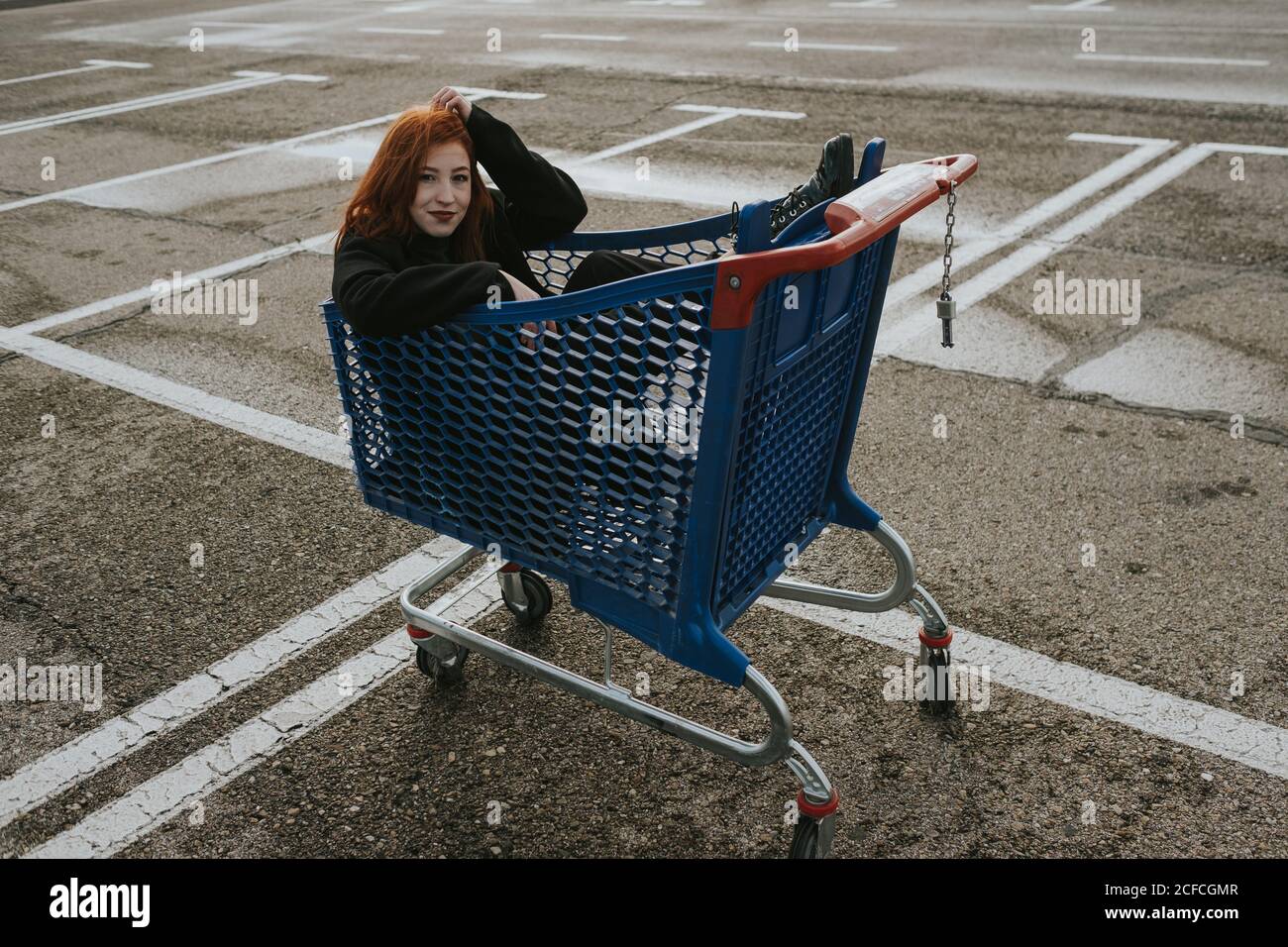 Mujer joven atractiva con pelo rojo en chaqueta negra teniendo diversión sentado en el carrito de compras en el estacionamiento marcado Foto de stock