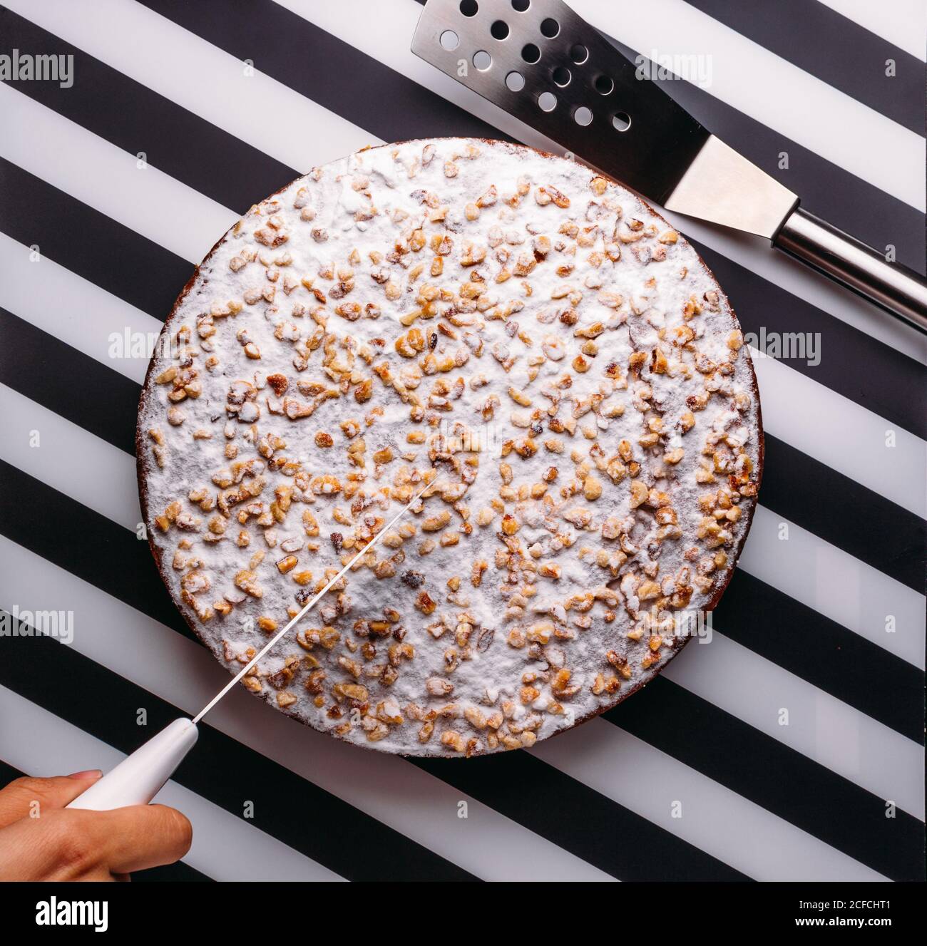 Vista superior de la persona de la cosecha cortando la torta entera con la formación de hielo azúcar y frutos secos en la superficie de rayas negras y blancas Foto de stock