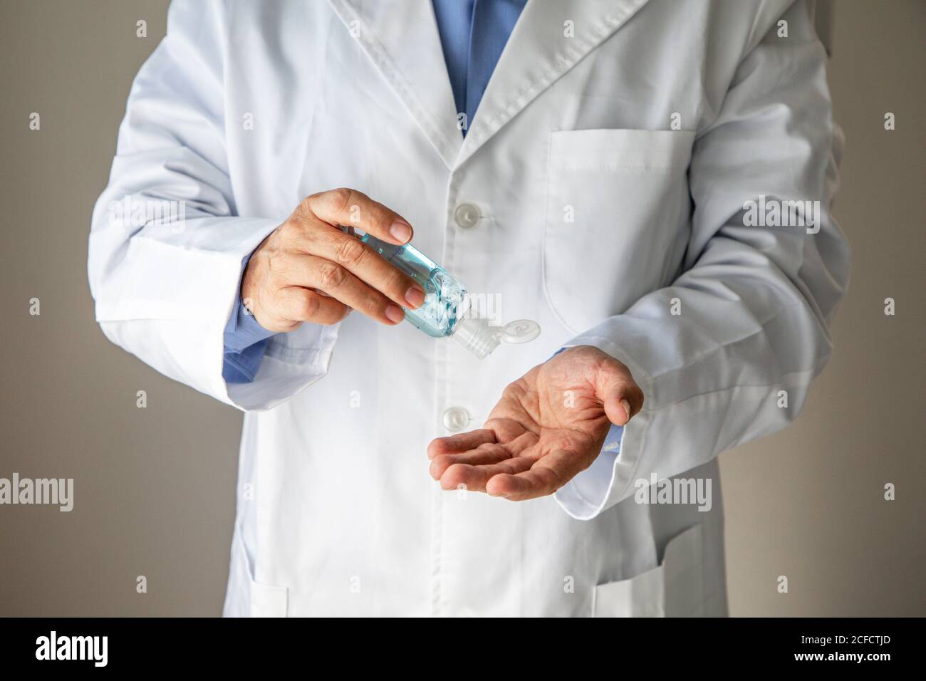 Crop masculino general practicante en bata médica aplicando gel antibacteriano para desinfectar las manos mientras está de pie sobre fondo gris en clínica durante la pandemia del coronavirus Foto de stock