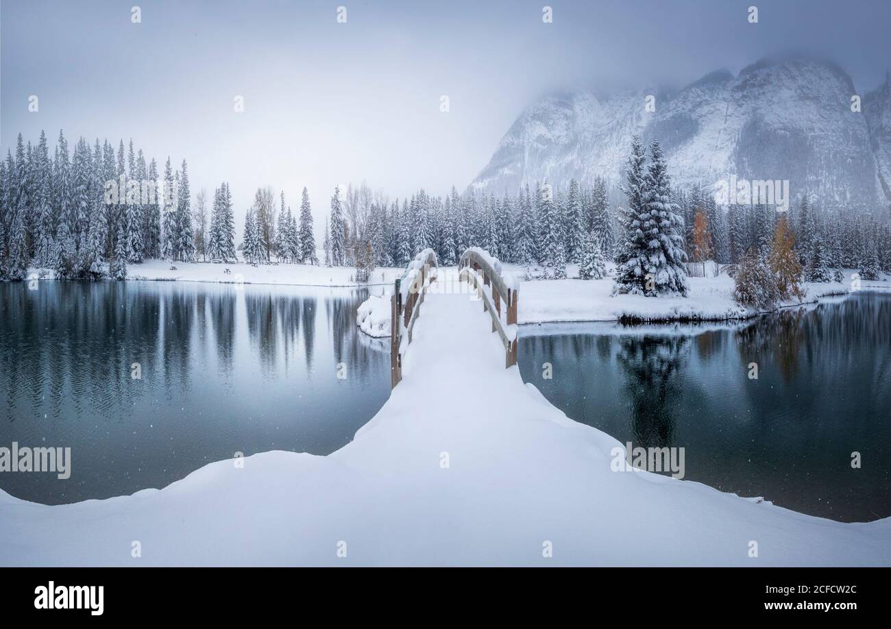 Hermoso paisaje canadiense de invierno con puente cubierto de nieve sobre la calma agua con bosque de abetos y montañas nevadas en el fondo Foto de stock