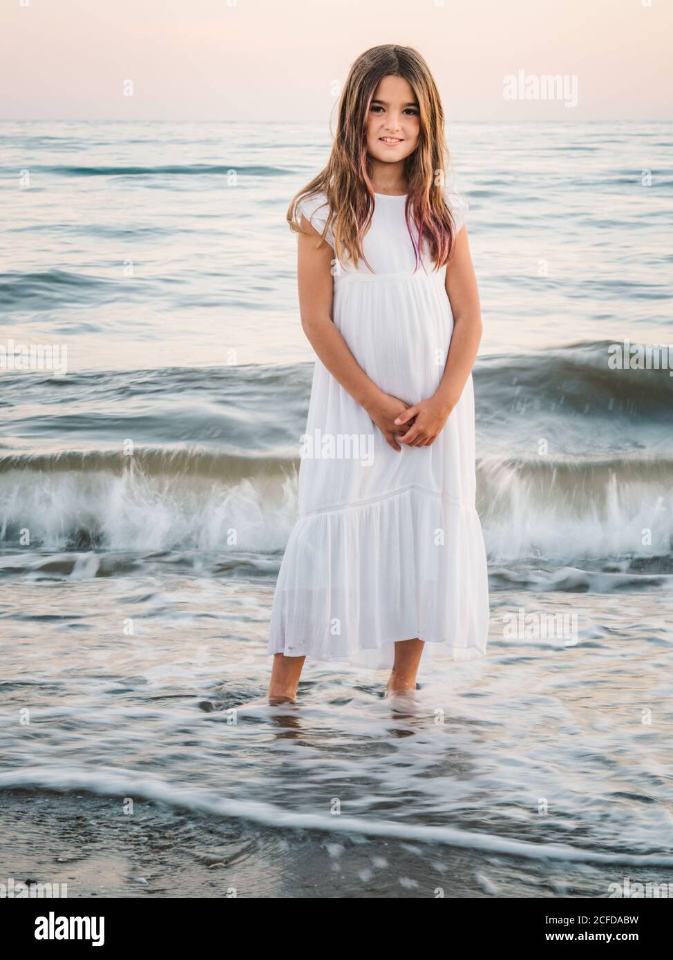 Retrato de niña encantadora con vestido blanco de pie agua en la playa de  arena y mirando la cámara Fotografía de stock - Alamy