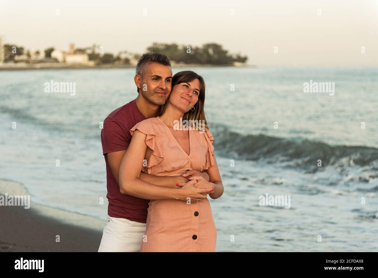 Hombre adulto abrazando a la mujer por detrás y mirando lejos mientras está de pie en la playa cerca del mar agitado y descansando juntos Foto de stock