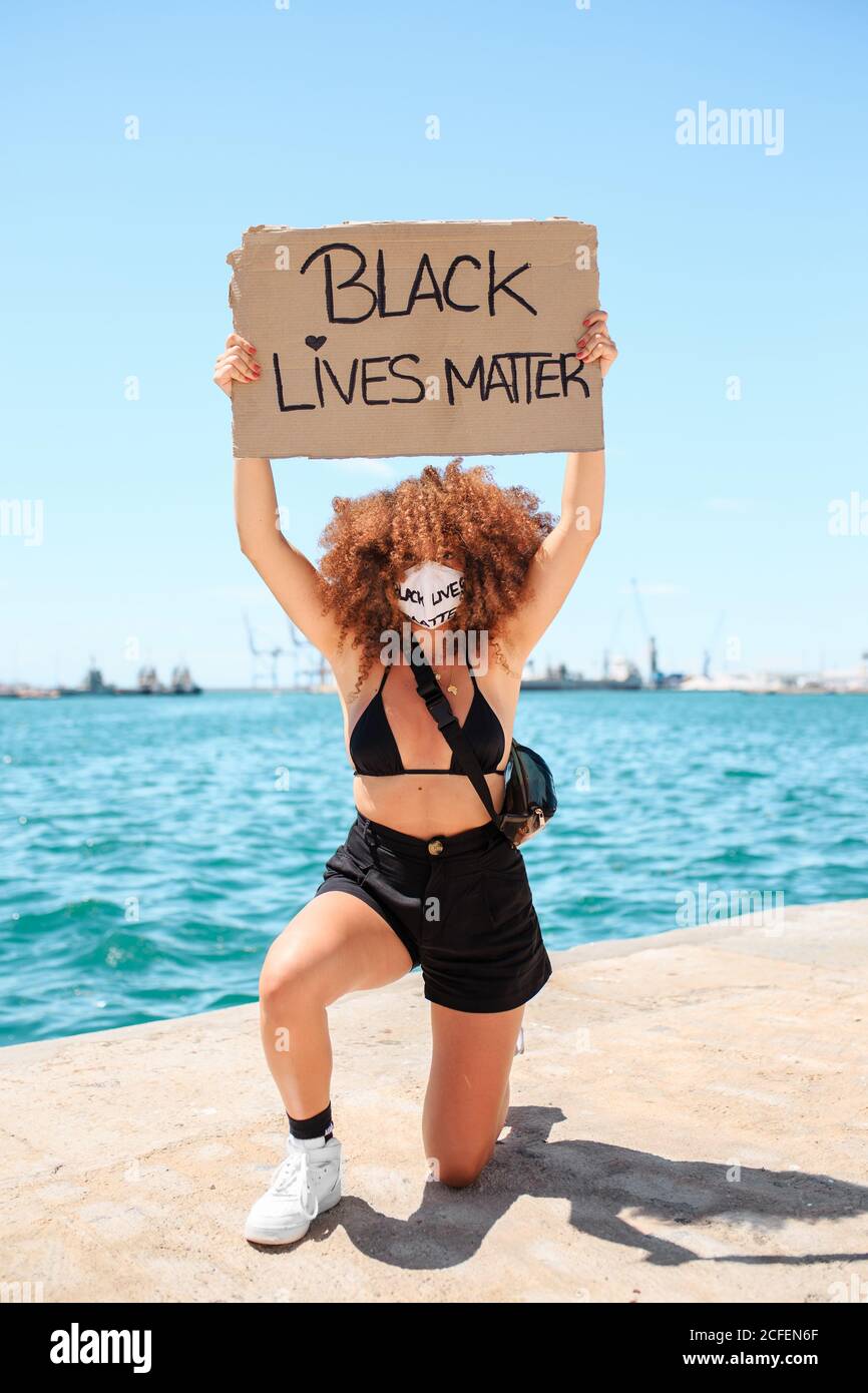 Mujer activista étnica en máscara médica y con vidas negras materia aplacard tomando la rodilla en solidaridad durante la protesta pacífica en la ciudad y mirando la cámara Foto de stock