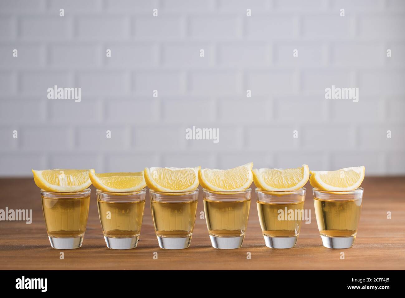 Fila de tequila de vidrio con tequila de oro y rebanadas de limón sobre mesa de madera con pared blanca sobre fondo borroso Foto de stock