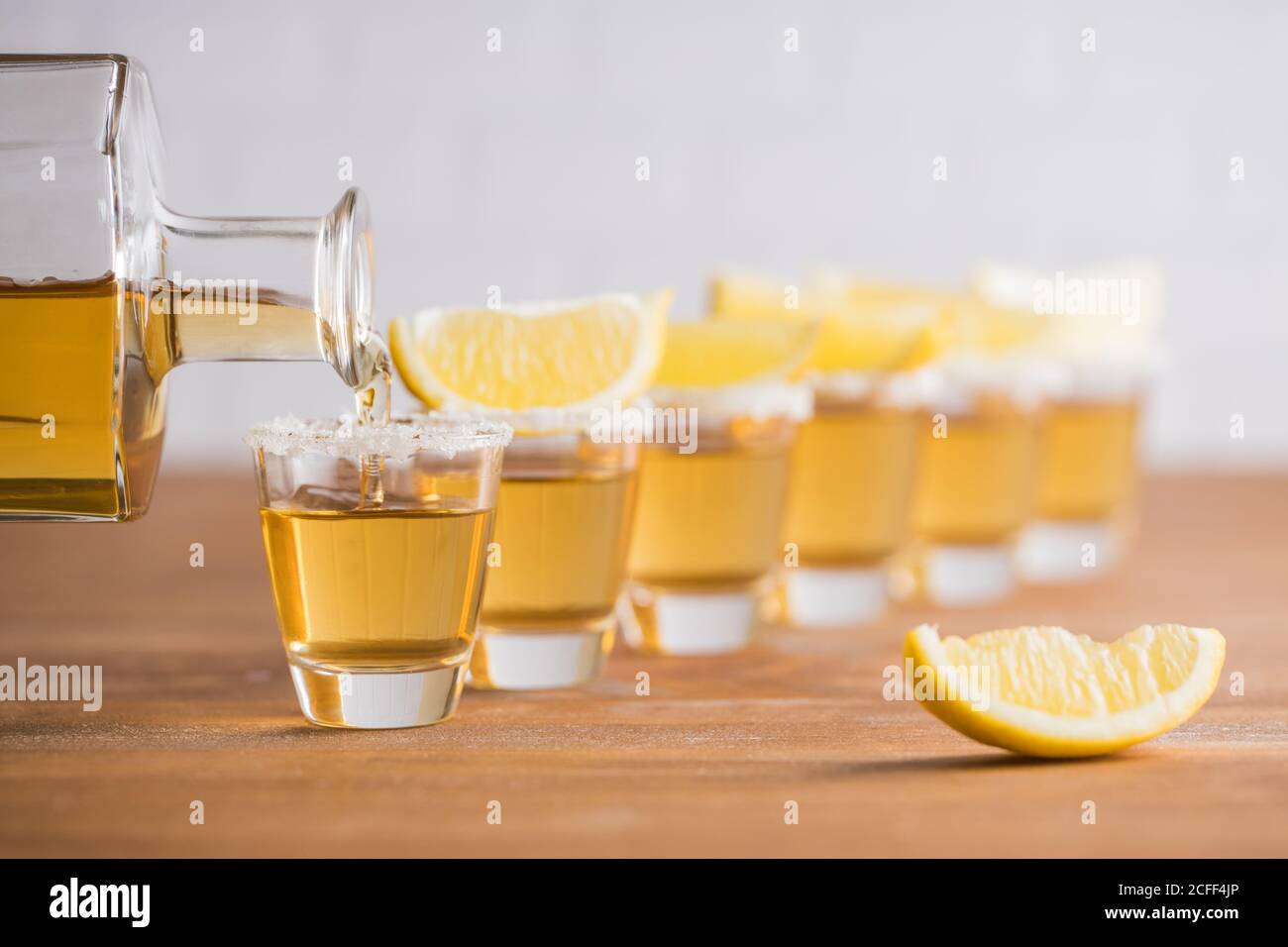 Fila de tequila de vidrio con tequila de oro y rebanadas de limón sobre mesa de madera con pared blanca sobre fondo borroso Foto de stock