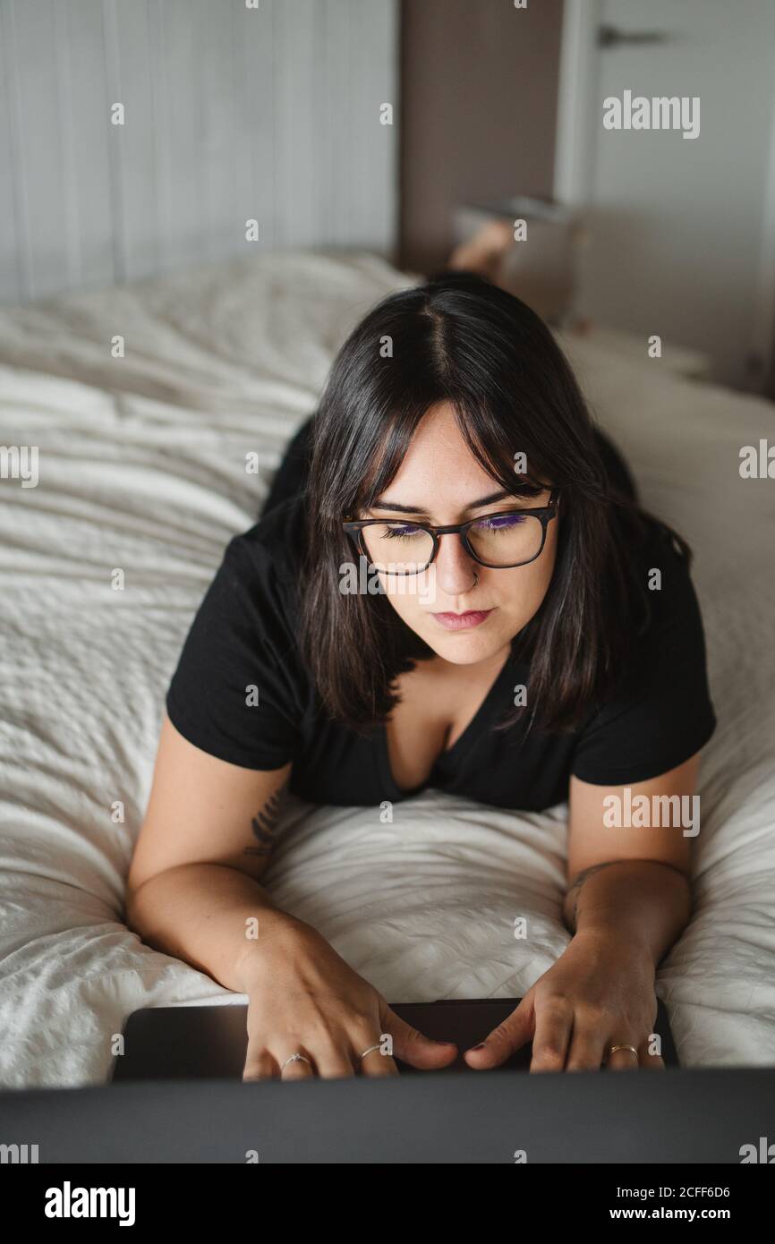 Desde la vista superior de la mujer joven concentrada en gafas mecanografiando en el ordenador portátil mientras estudia y se acuesta en la cama en casa en París Foto de stock