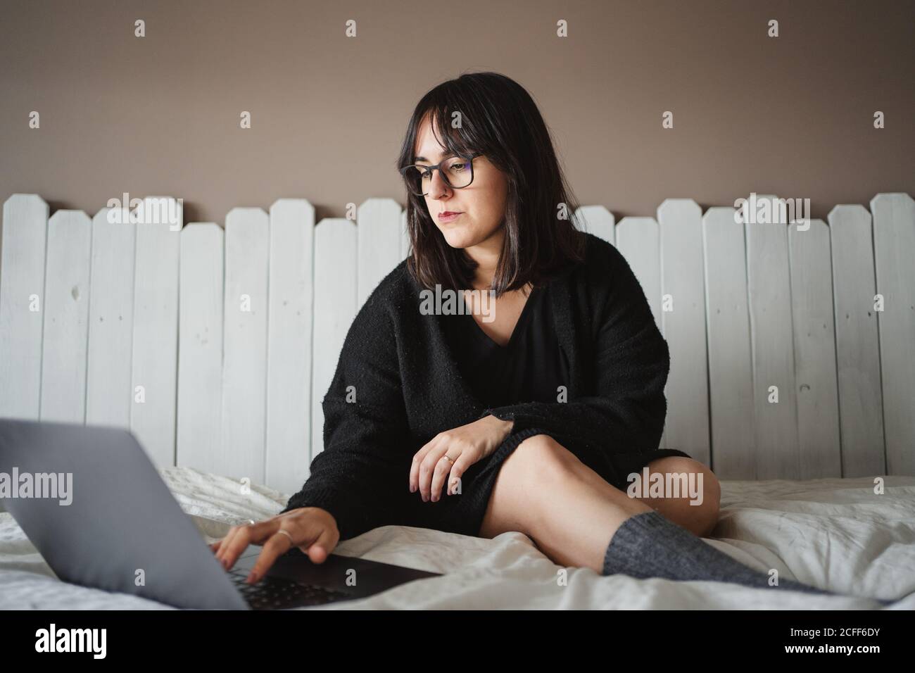 Vista de la mujer joven concentrada en gafas mecanografiando en el ordenador portátil mientras estudia y se acuesta en la cama en casa en París Foto de stock