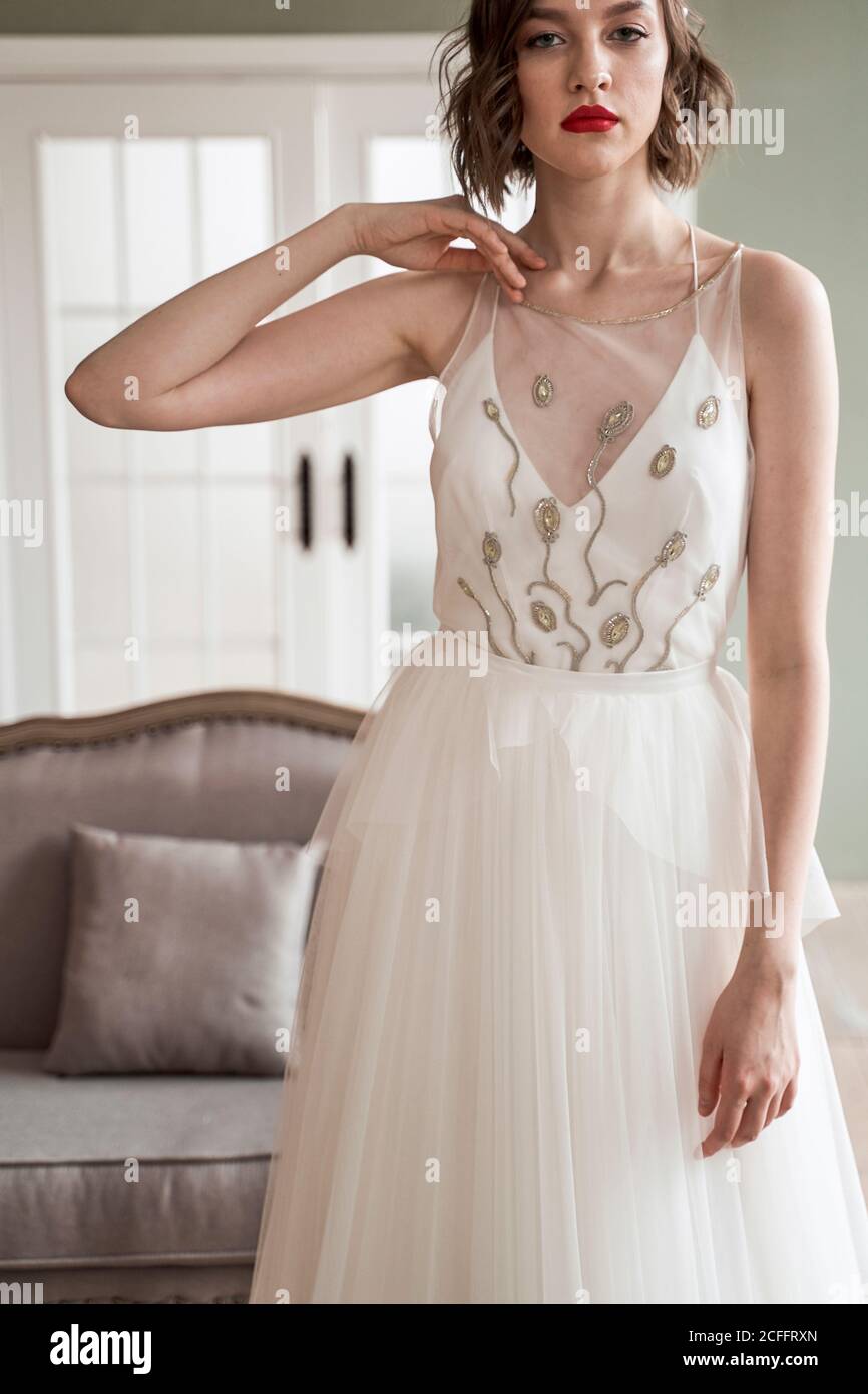 Mujer de la cosecha en vestido de boda de moda con tela transparente y..  patrón dorado mirando la cámara mientras se está de pie y tocando el cuello  contra el interior borroso