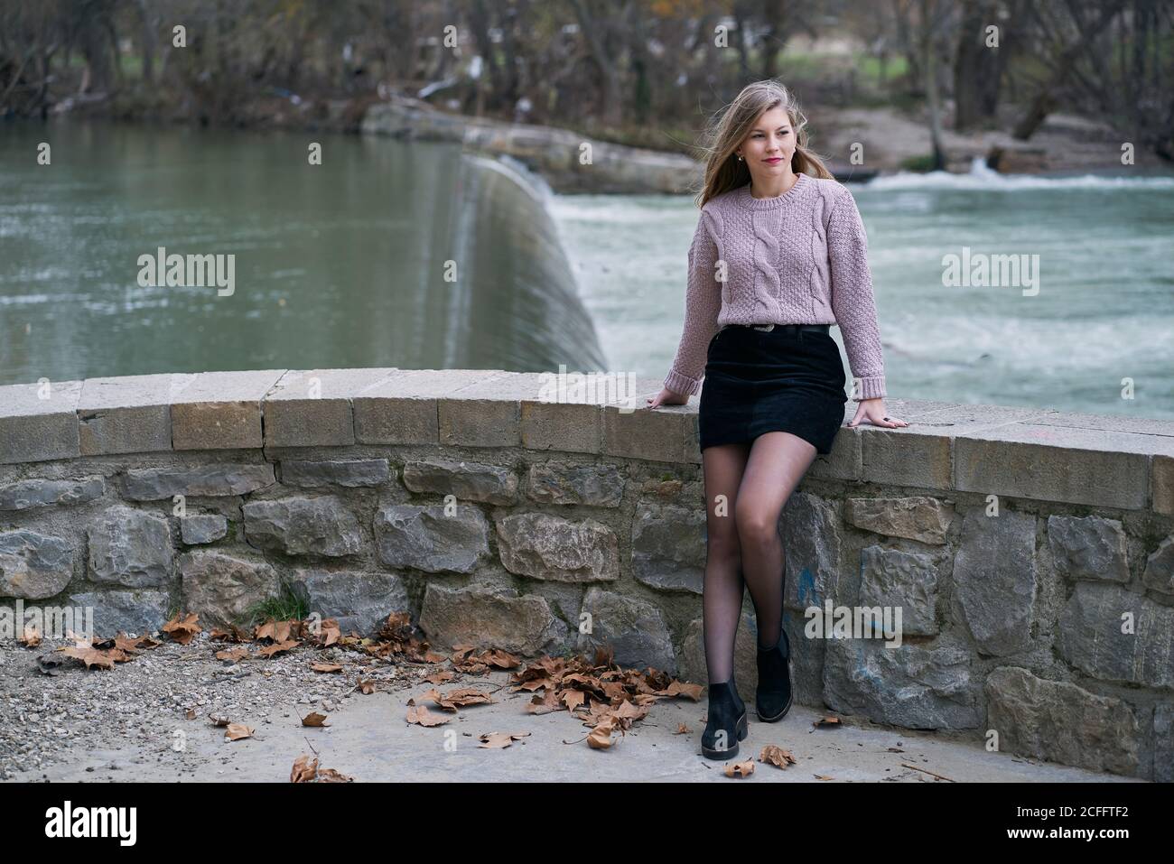 Mujer tranquila con suéter de punto y falda negra corta y apoyándose en una valla de piedra mientras caminaba cerca del río Fotografía de stock Alamy