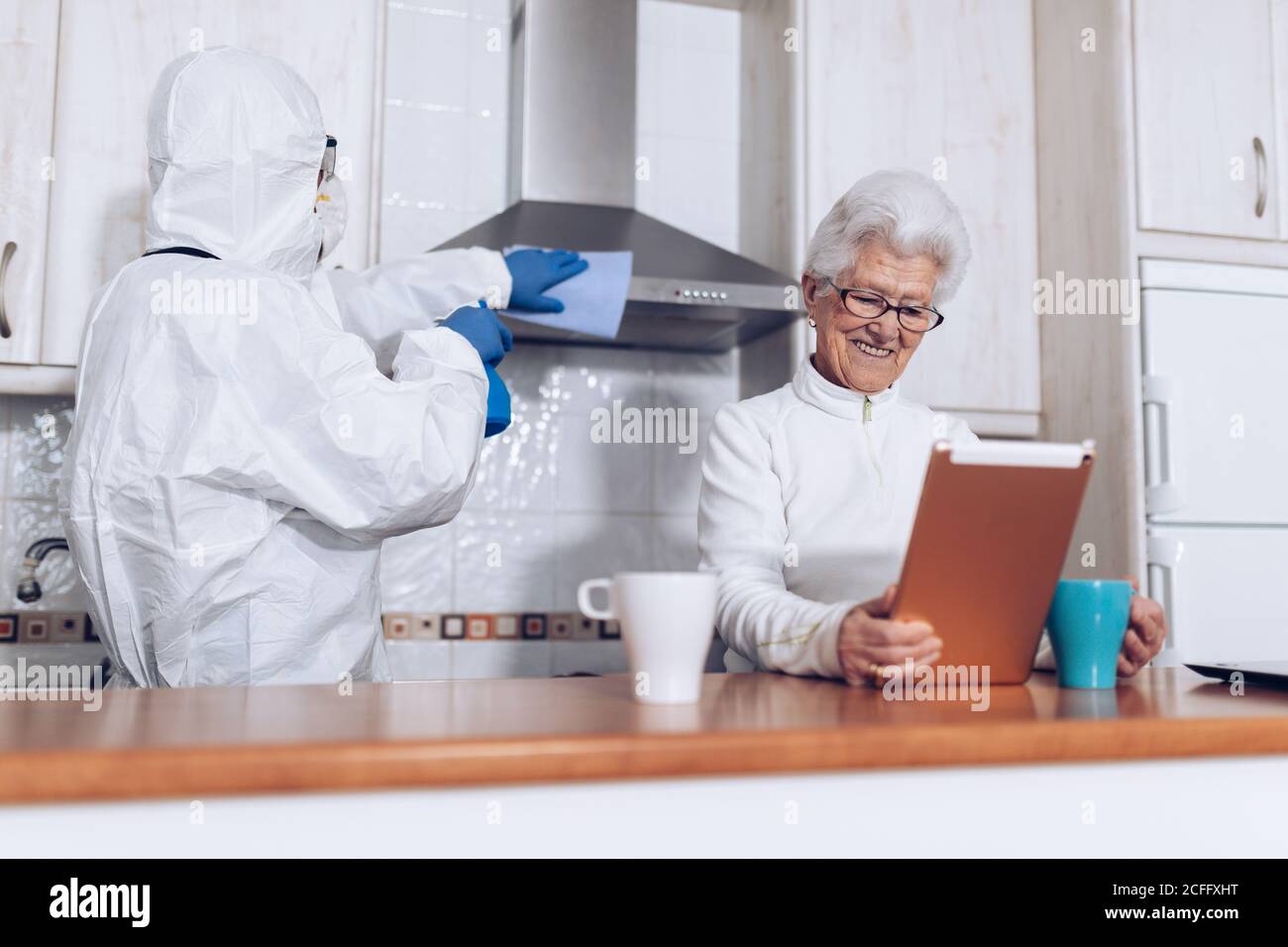 Irreconocible trabajador de atención domiciliaria en la cocina de limpieza uniforme de protección mientras alegre mujer anciana disfrutando de nuevo libro durante el autoaislamiento en casa debido a la pandemia de coronavirus Foto de stock