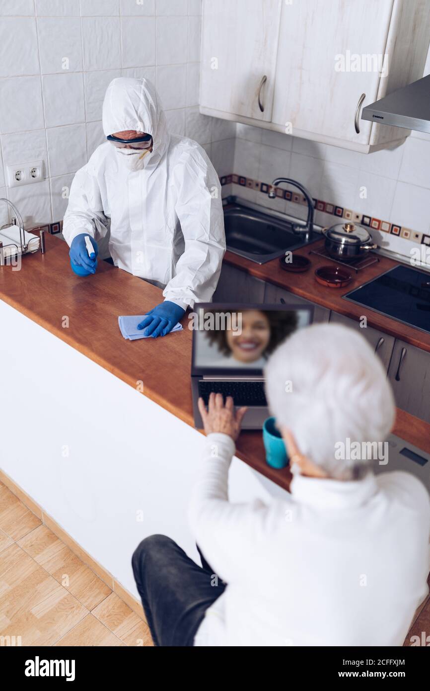 Mujer anciana charlando en el ordenador portátil y un empleado de atención domiciliaria desinfectando la cocina durante una pandemia Foto de stock