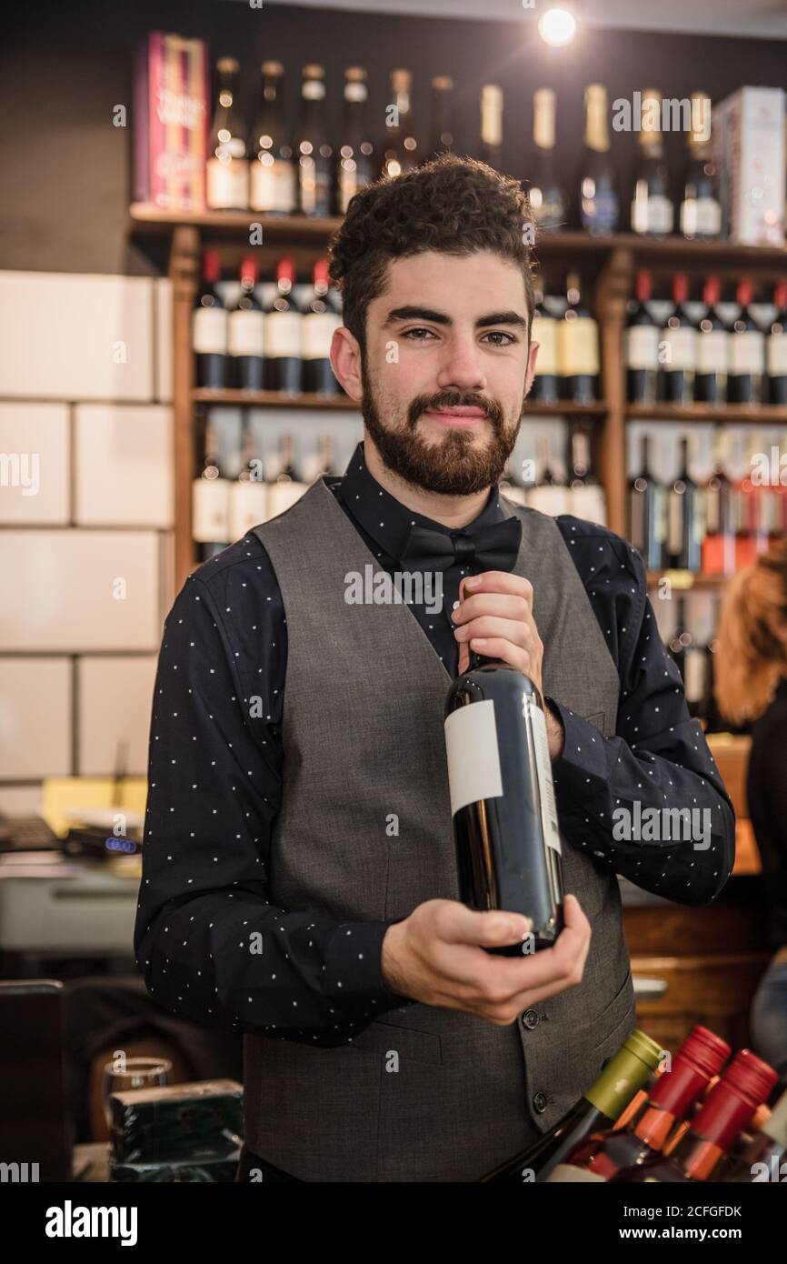 Hombre adulto mirando la cámara mientras sostiene una botella de vino en la tienda de vinos. Foto de stock