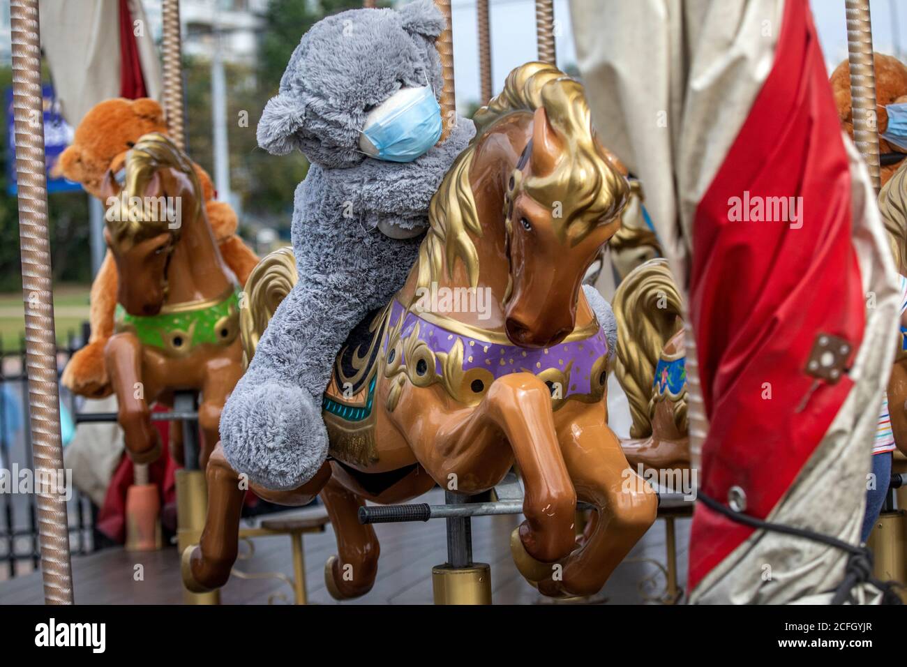 Un juguete de oso de peluche con una máscara protectora se asienta un caballo de carrusel en una ciudad durante las restricciones con La nueva pandemia de coronavirus COIVD-19 Foto de stock