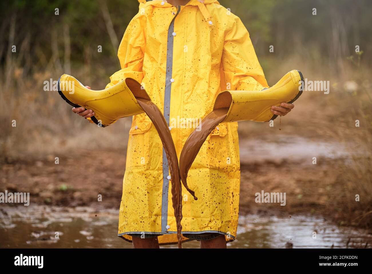 Persona con impermeable amarillo jugando con las botas de agua llenarlos con agua de un charco y vaciarlos en medio del bosque Foto de stock