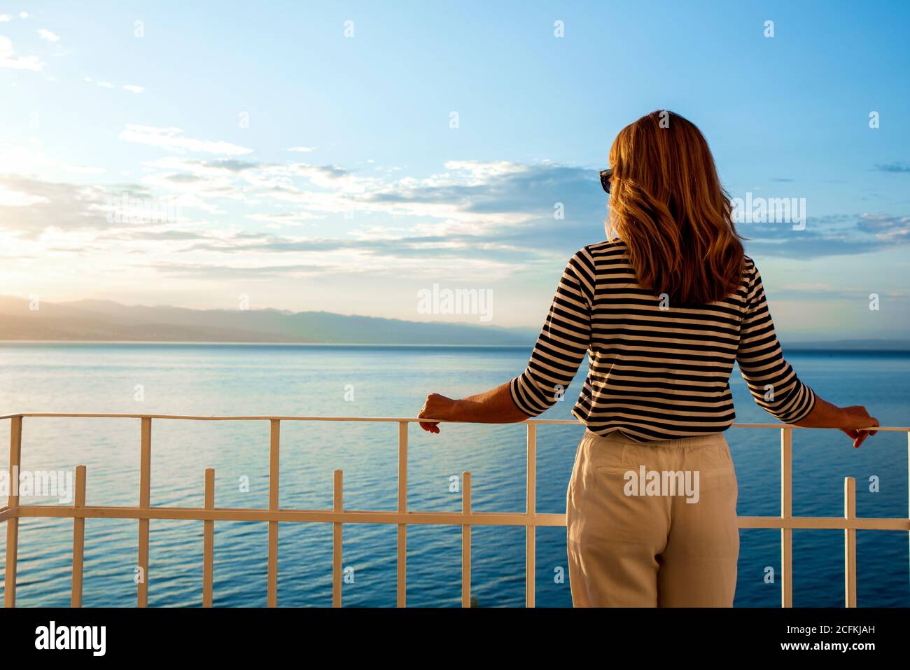 Un tiro de mujer bonita con sombrero de paja y gafas de sol mientras estaba de pie en el balcón y mirando a la vista al mar. Soñando despierto. Foto de stock