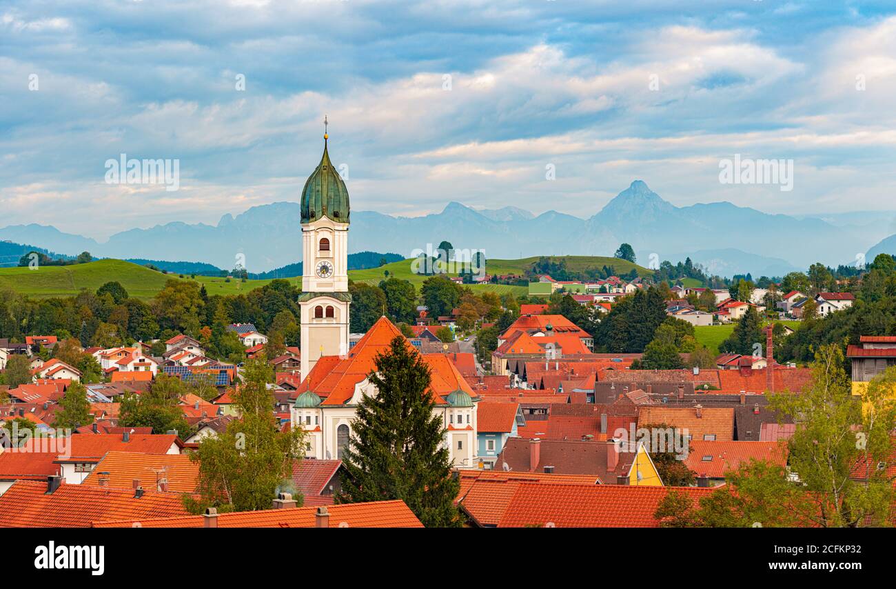 Hermoso y acogedor pequeño pueblo bávaro en Alemania, Europa. Iglesia y casas en primer plano con cielo nublado y montañas en el fondo Foto de stock
