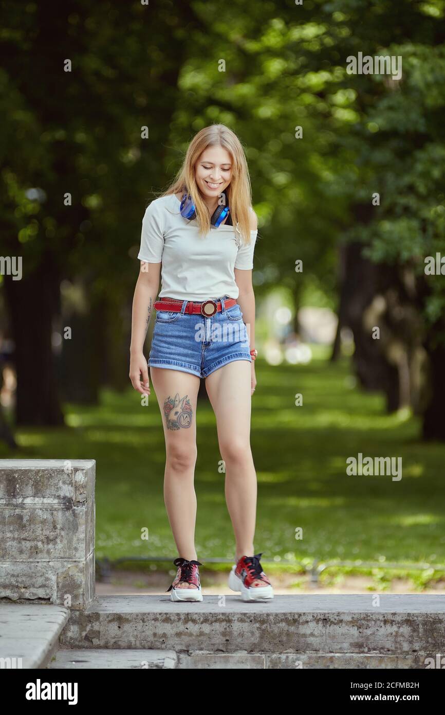 Una mujer rusa sonriente en pantalones cortos vaqueros tiene un descanso en  el parque público durante el día de verano. La niña de 20 años de edad en  tela de moda está