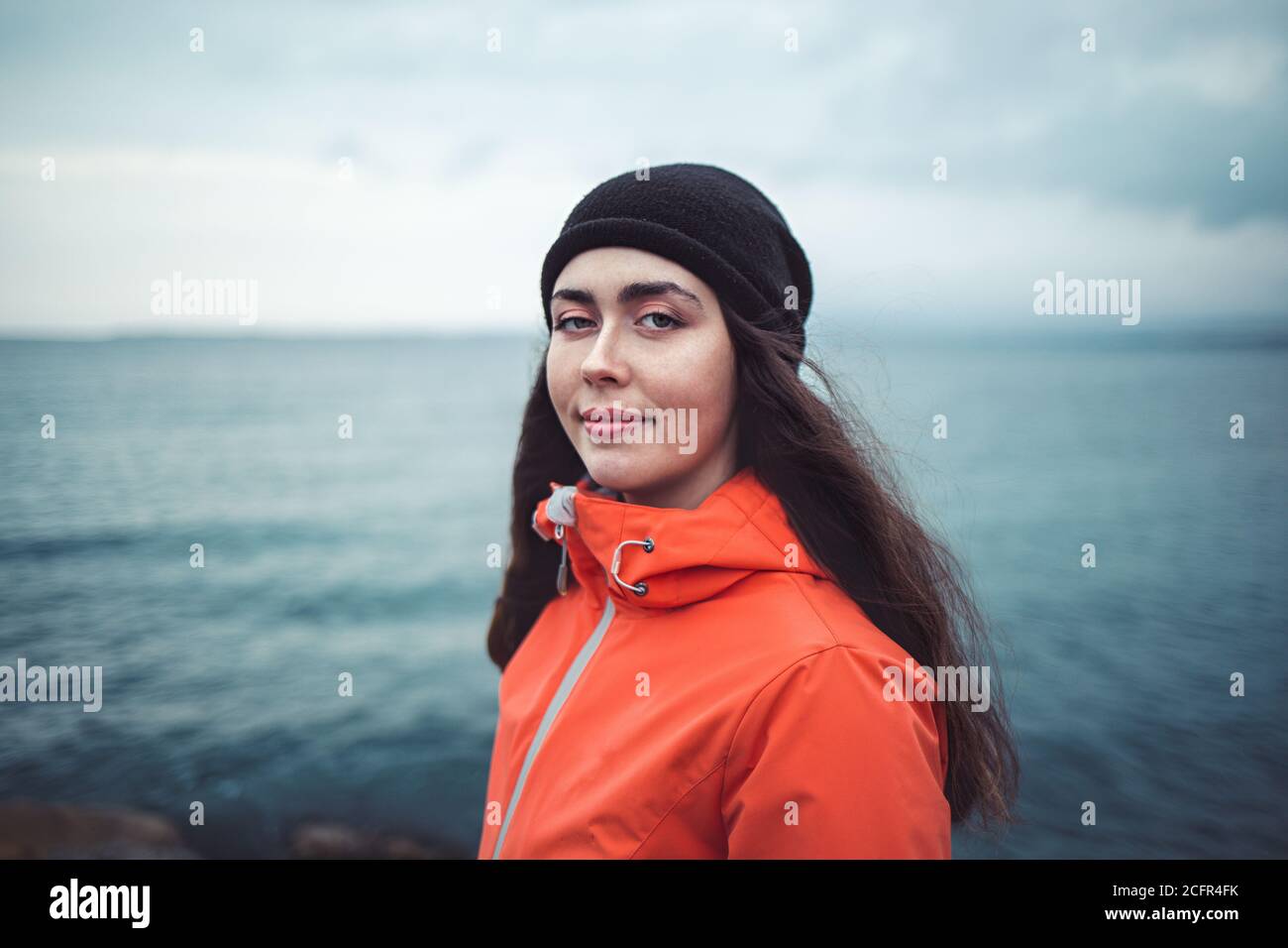 Retrato de una mujer morena sonriente con pelo largo, con un sombrero y una chaqueta naranja. En el fondo el mar y la línea del horizonte. Espacio de copia. Foto de stock