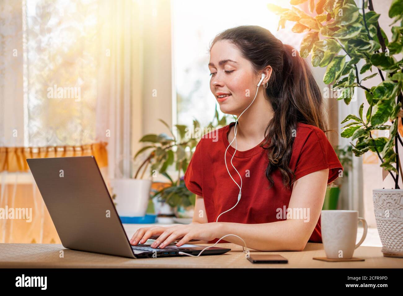 Freelance.UNA joven mujer caucásica feliz escribiendo en un ordenador portátil con auriculares en sus oídos.Home environment.Concept de cuarentena, teletrabajo y dista Foto de stock