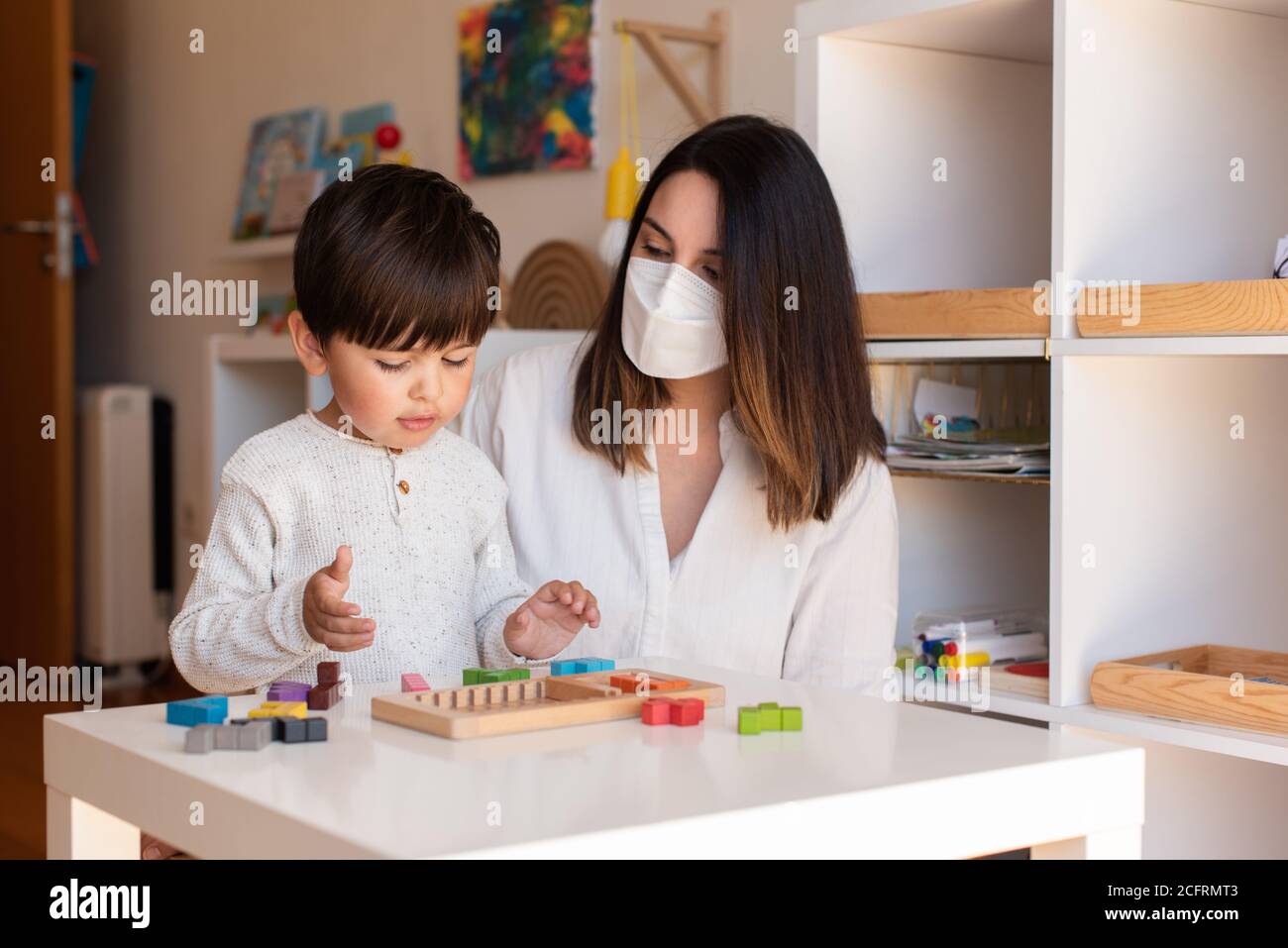 Liltle Preschooler Kid jugando al rompecabezas de madera de tetris con el maestro educador ayuda a usar la máscara para la pandemia del coronavirus. Resolución de problemas en casa. Unidad de aprendizaje Foto de stock