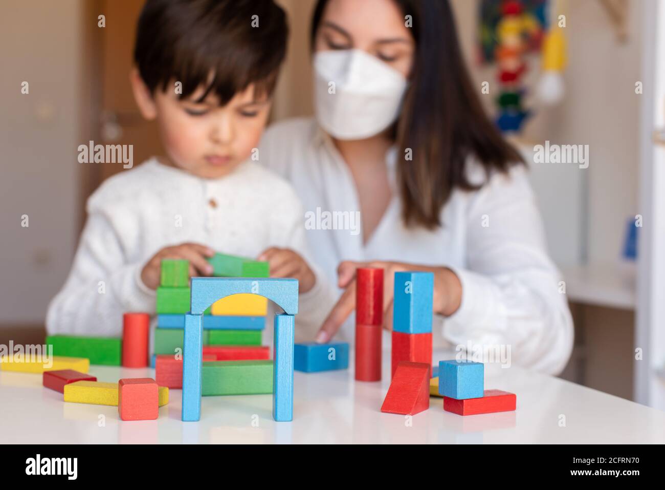 Liltle Preschooler Kid jugando con bloques de madera y maestro educador ayuda a usar la máscara para la pandemia del coronavirus. Resolución de problemas en casa. Comunidad de aprendizaje. Foto de stock