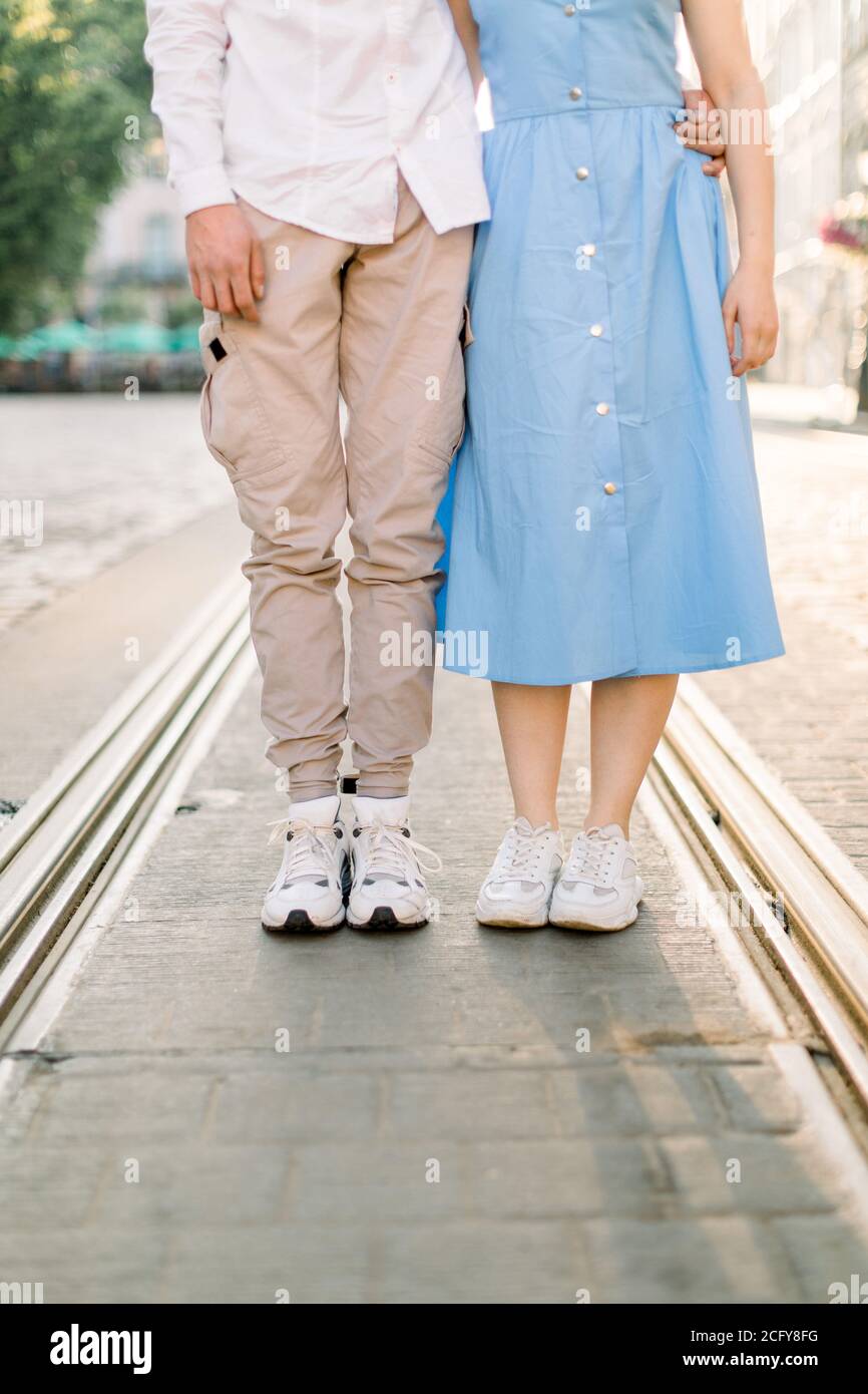 Imagen de las piernas de pareja joven y con estilo enamorada, con vestido azul y hombre con pantalones beige, de pie en el camino del pavimento y la
