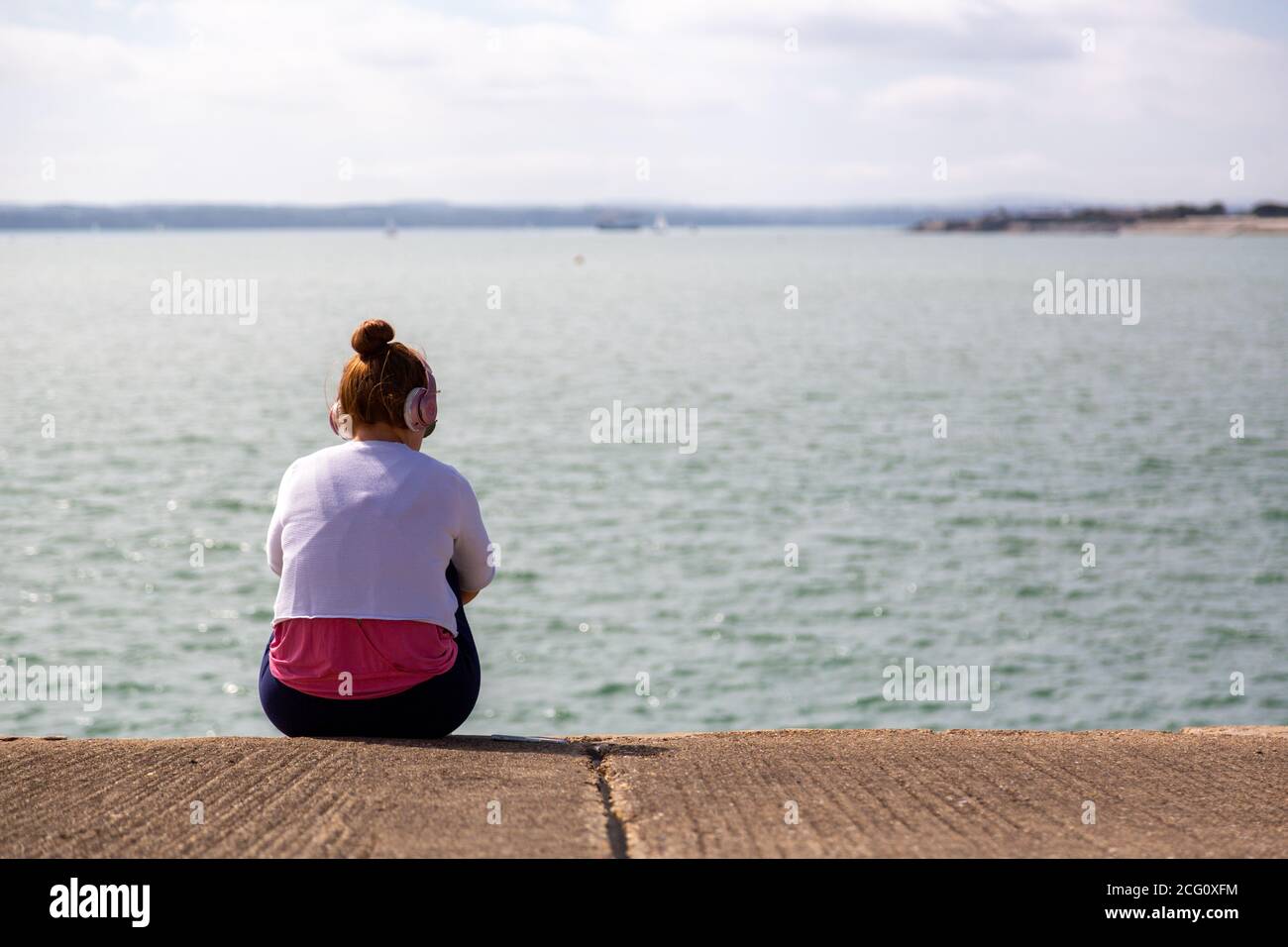 Un adolescente sentado solo en una pared escuchando música en los auriculares mirando una vista del mar Foto de stock