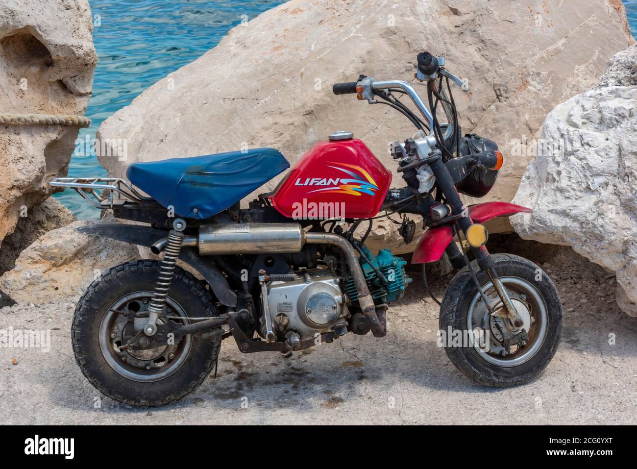 una mini moto con un tanque de gasolina rojo apoyado sobre algunas rocas en la costa de grecia. Foto de stock