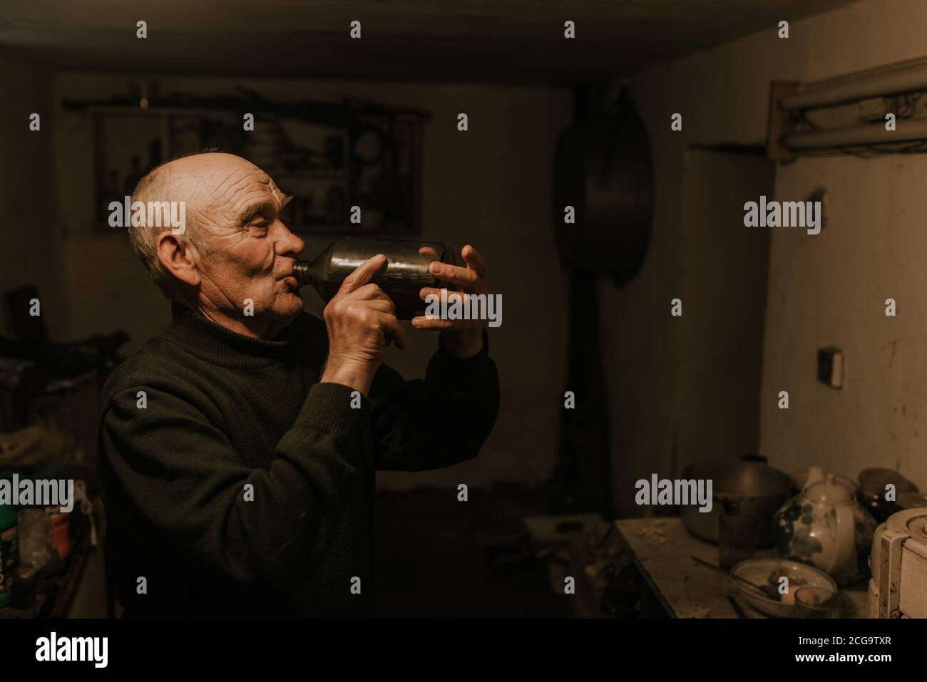 el viejo hombre de pelo gris con arrugas profundas bebe vino tinto casero de una botella de vidrio en el oscuro sótano de la casa. Foto de stock