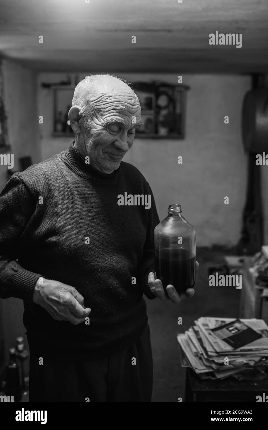 el viejo hombre de pelo gris con arrugas profundas bebe vino tinto casero de una botella de vidrio en el oscuro sótano de la casa. Foto de stock