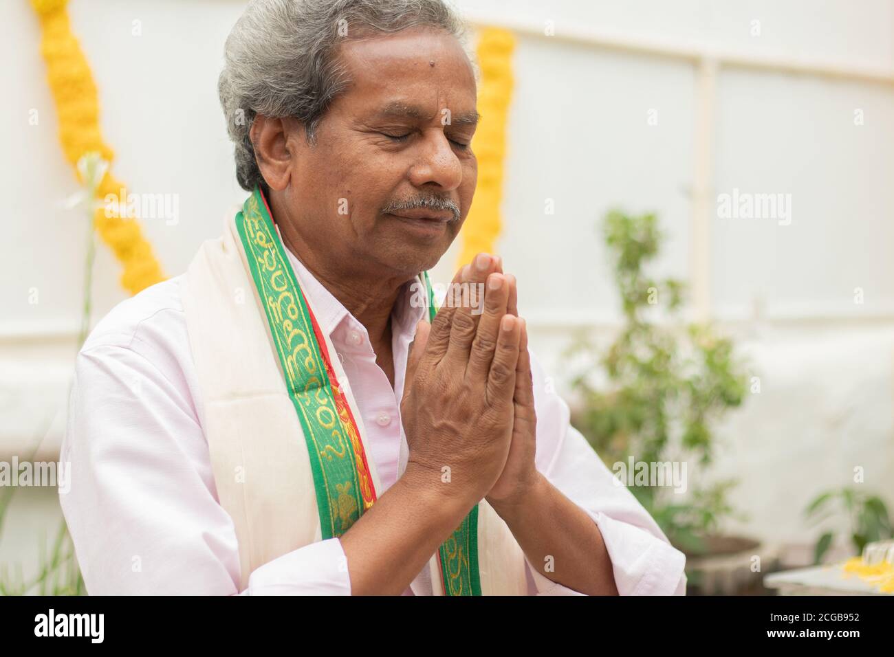 Anciano a ojos cerrados ofreciendo oración por gesto de namaste rezando a dios en casa con vestimenta tradicional durante los rituales. Foto de stock