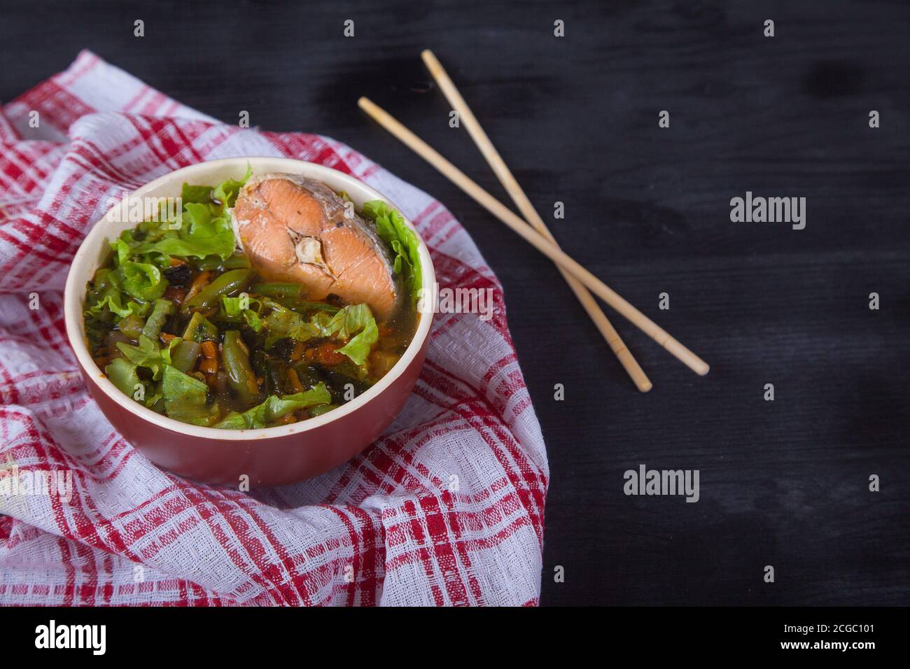 Sopa japonesa con salmón, judías verdes, hierbas y palillos de bambú sobre una servilleta roja sobre fondo negro. Copiar espas Foto de stock