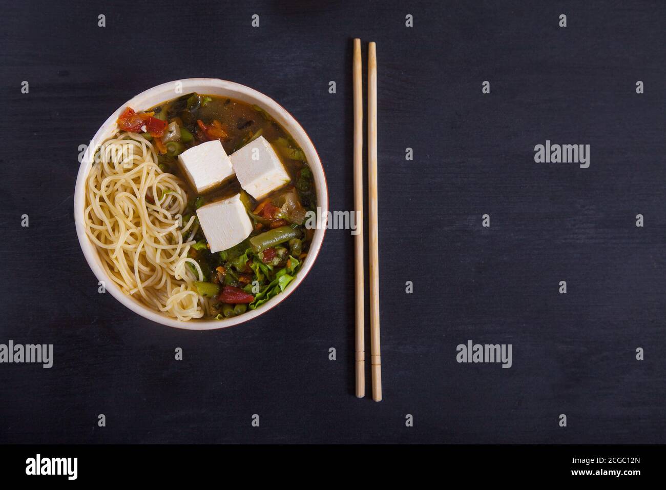 Sopa japonesa con tofu, fideos, verduras y hierbas, palillos de bambú sobre fondo negro. Copiar espas Foto de stock