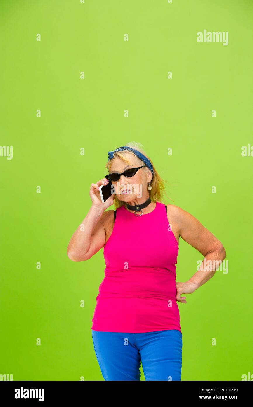 Hablando por teléfono. Mujer mayor en traje ultra aislado sobre fondo verde brillante. Se ve con estilo y de moda, para siempre joven. Mujer caucásica madura gafas de sol, ropa